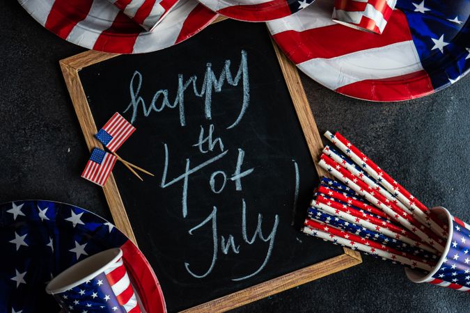 Chalkboard surrounded by USA flag plates, cups and straws, with the words "Happy 4th of July"