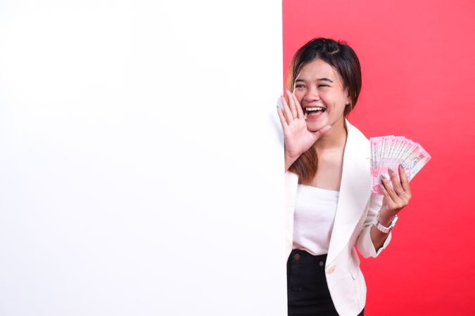 A woman is holding paper money and shouting with her hand near her mouth in a jacket and pants, isolated on a red background