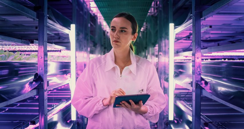 Portrait of Female Agricultural Engineer Walking in Vertical Farm Facility Next to Racks with Fresh Natural Crops. Professional Hydroponics Specialist Using Tablet, Studying, Cultivating Plants.