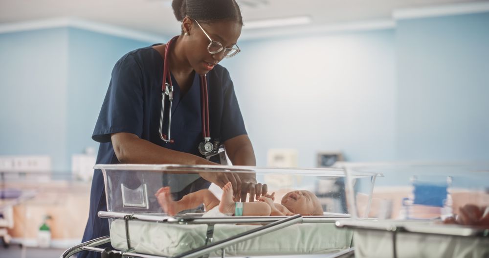 Newborn Baby Lying in Bassinet in a Maternity Hospital With Nurse Attending