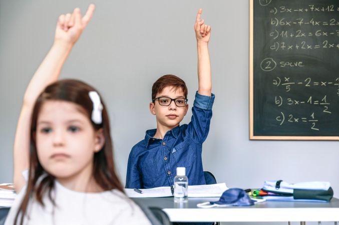 Students with mask on table raising hands at school