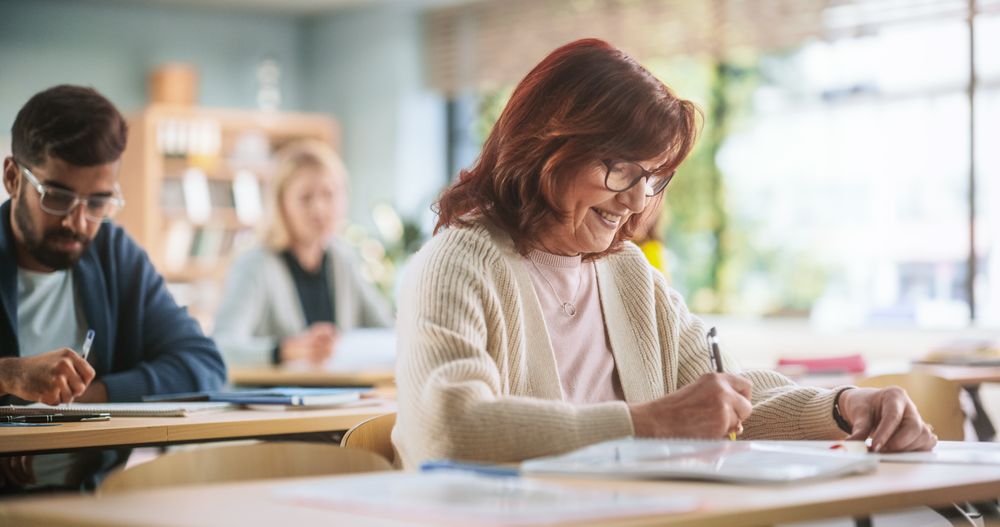 Older Female Wearing Glasses, Sitting Behind a Desk, Using Textbook, and Writing Down Notes in Notebook