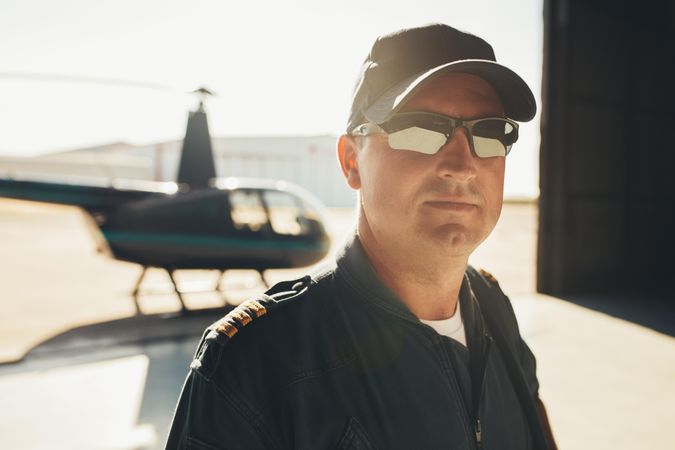 Pilot in cap and sunglasses inside hangar with helicopter in background