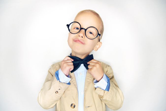 Blond boy making an inquisitive face wearing round glasses and fixing his bow tie