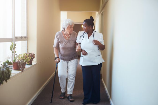 Older woman with walking stick being helped by a female nurse at home