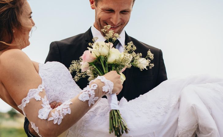 Happy groom carrying beautiful bride with flowers in hand