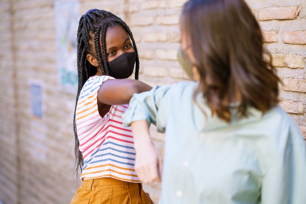 Female friends tapping elbows in lieu of shaking hands in front of brick wall