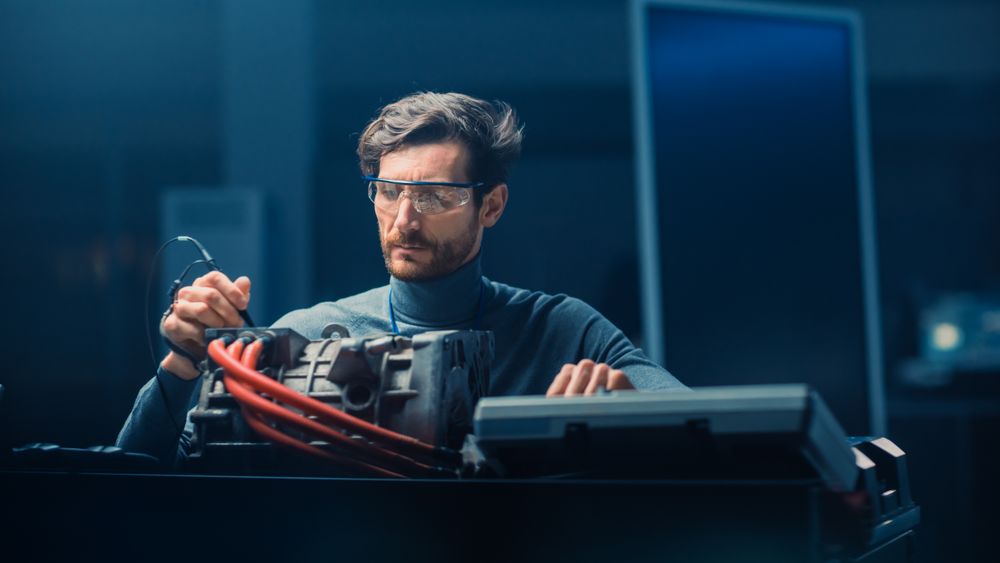 Professional Automotive Engineer in Glasses with a Computer and Inspection Tools is Testing an Used Electric Engine in a High Tech Laboratory with a Concept Car Chassis.