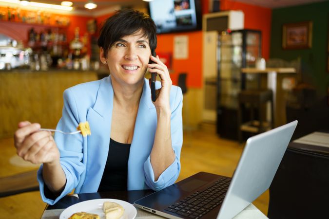 Happy woman sitting in cafe speaking on phone with laptop and slice of cake