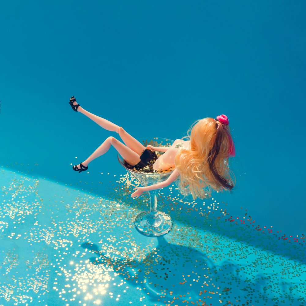 Doll bathing in martini glass full of gold glitter on blue background
