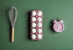 Cooking concept with pink egg box, whisk, and alarm clock