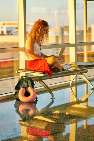 Enthusiastic woman working on laptop sitting on bench in airport hall