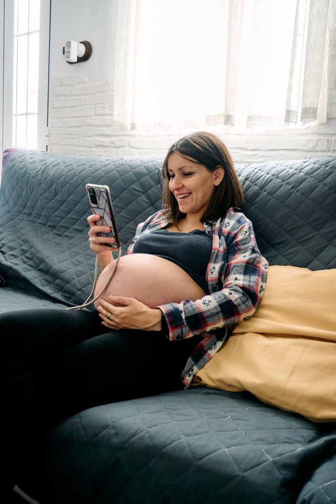 A young woman with a very advanced pregnancy is sitting on the couch looking at and talking on her cell phone.