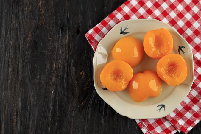 Plate of tinned peaches on dark wooden table with checkered napkin