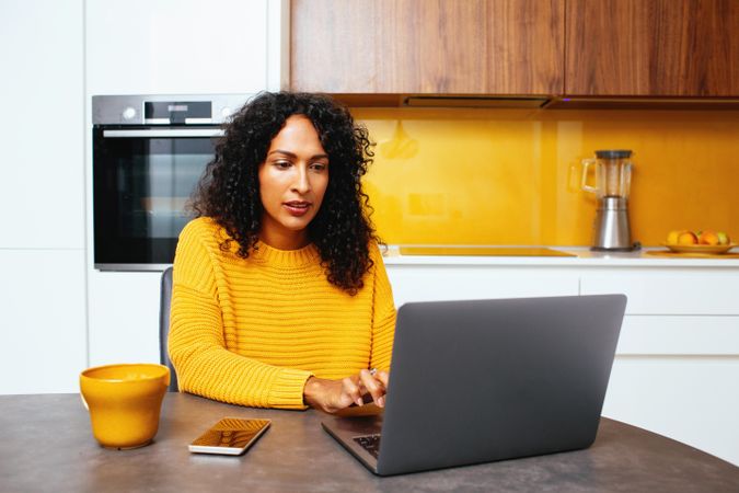 Woman in colorful kitchen smiling concentrating on her laptop with mug and smart phone
