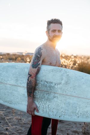 Tattooed man smiling with surfboard under his arm with sun at his back