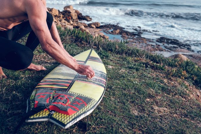 Crop male surfer rubbing surfboard with wax on rocky seashore