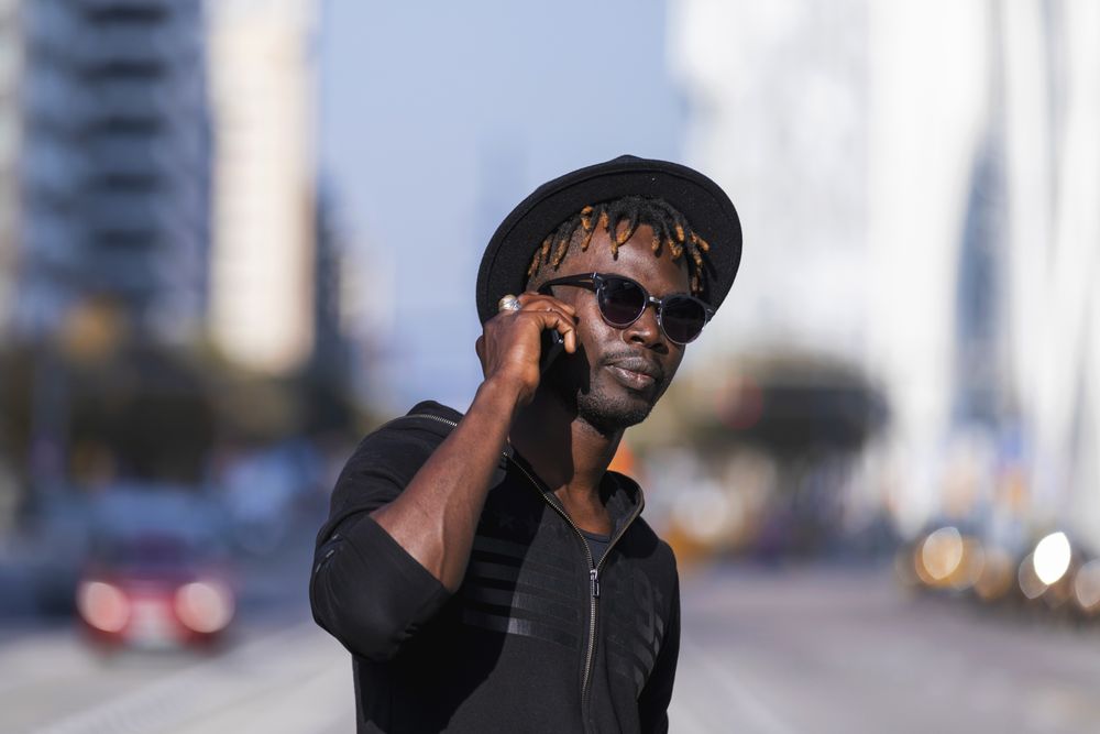 Black man wearing hat & sunglasses standing on the street talking on cellphone