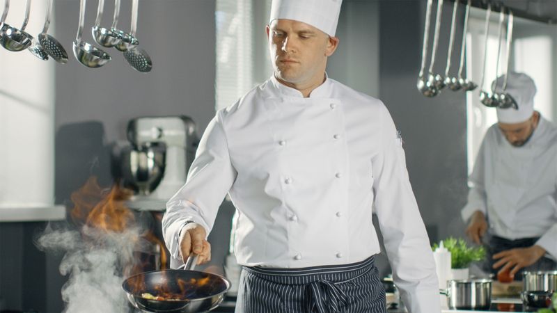 Professional Chef Fires up Oil on a Pan. Flambe Style Cooking. He Works in a Modern Kitchen with Lots of Ingredients are Around.