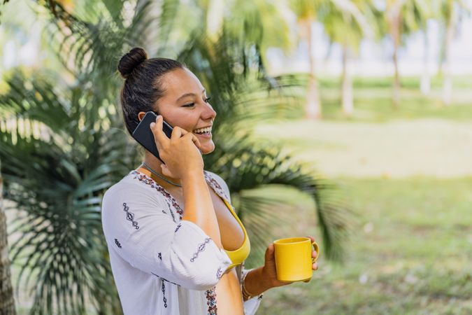 Cheerful woman talking on phone while drinking coffee by palm tree