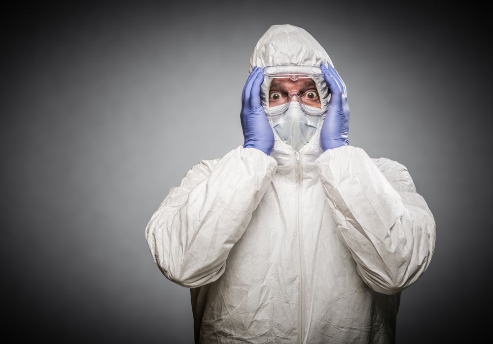 Man Holding Head With Hands Wearing HAZMAT Protective Clothing Against A Gray Background.