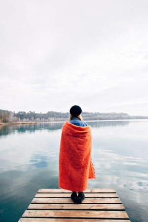 Woman wrapped in blanket looking out at lake from the pier