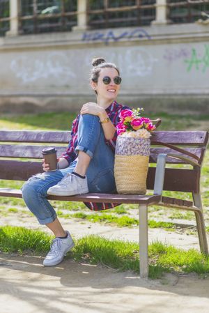 Front view of a young hipster woman sitting on a park bench relaxing in a sunny day while looking away