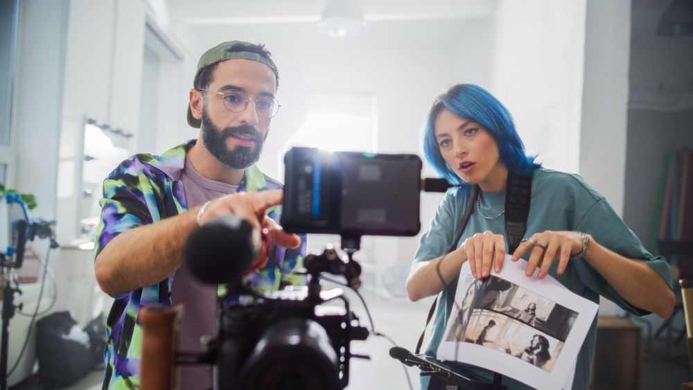 In A Bright Studio, A Young Male And Female With Blue Hair Review Footage On A Digital Camera, Surrounded By Filmmaking Equipment, Embodying Creativity And Collaboration In Movie Production.