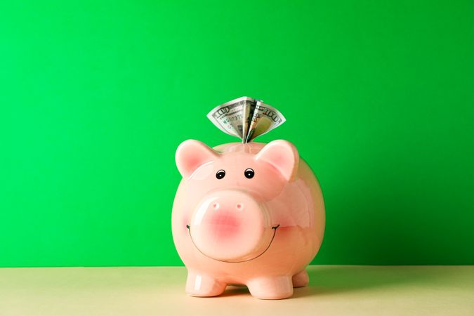 Happy piggy bank with one hundred dollar on color table against green background, space for text. Finance, saving money