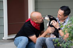 Man holding his dog and his partner leaning in giving the pet affection