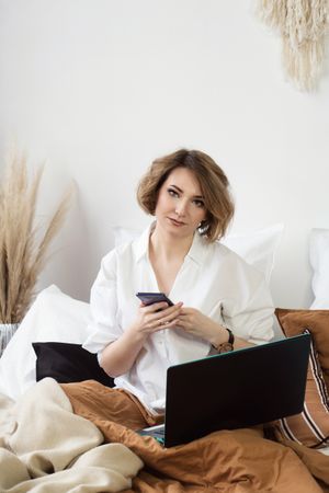 Woman Sitting In Bed Spending Time In Bedroom With Phone And Laptop