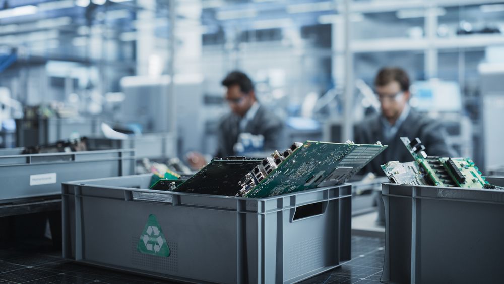 Indian And White Male Workers Taking Apart Laptops To Recycle For Production Of Digital Devices At Automated Electronics Factory With Robotic Arms.