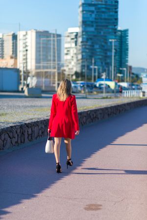 Rear image of a blonde woman wearing red jacket and holding a handbag while walking in city