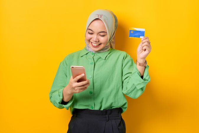 Muslim woman in headscarf and green blouse holding credit card and looking down at her smart phone