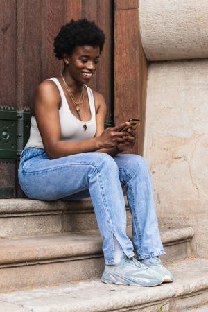 Smiling Black woman using smartphone sitting near building