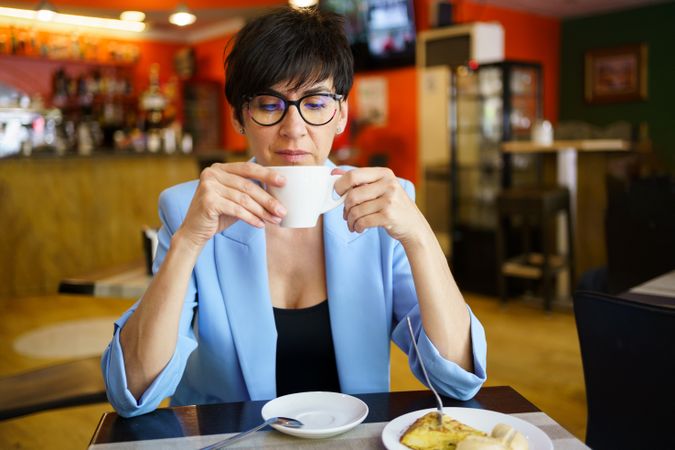 Positive female in trendy blue jacket and eyeglasses sitting at table with cup of hot drink and looking down