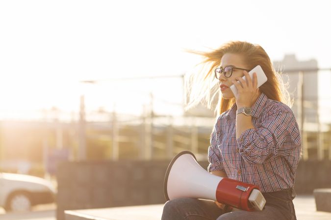 Woman sitting outside speaking on phone with megaphone dressed in checkered shirt