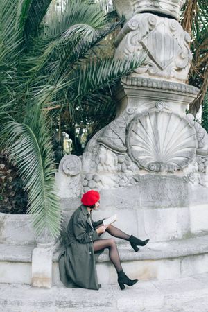 Side view of woman in green coat with red beret sitting beside statue outdoor in Paris