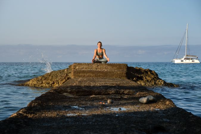 Woman practicing yoga while sitting on boulder near blue ocean