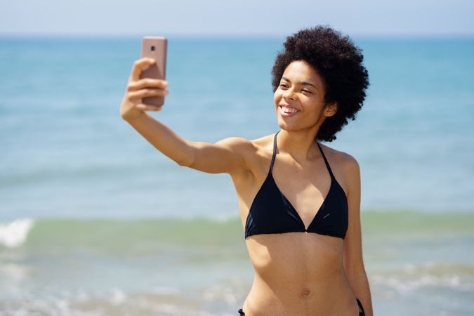 Joyful woman in bikini taking selfie on phone on coast