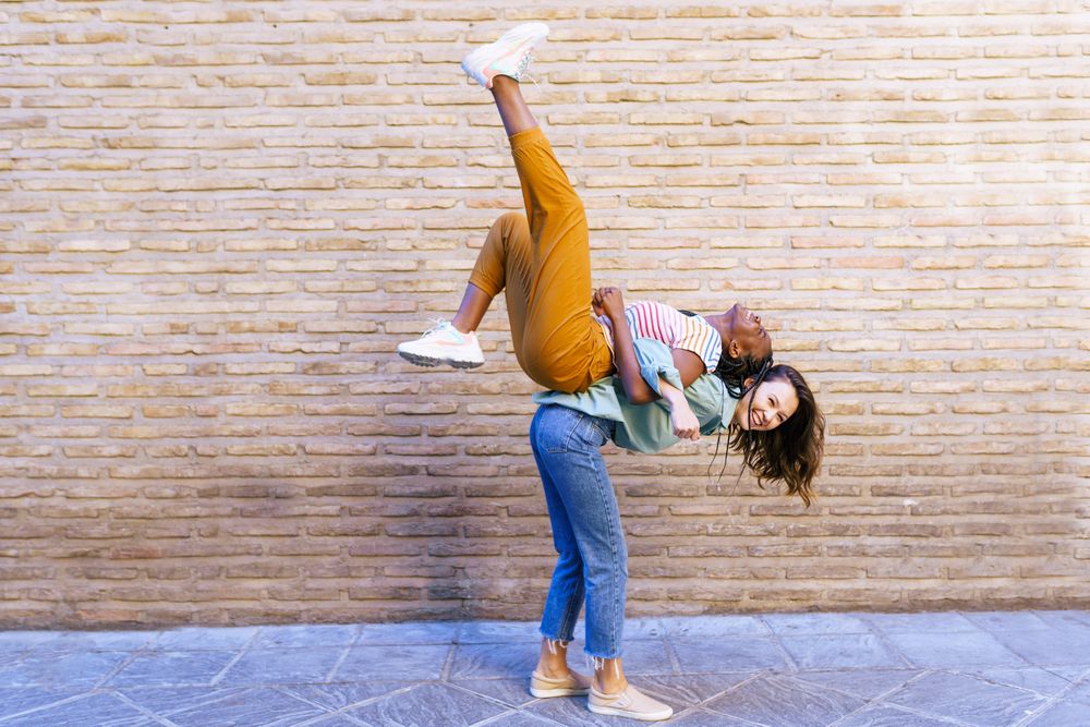 Two good female friends with one lifting the other up in front of brick wall outside