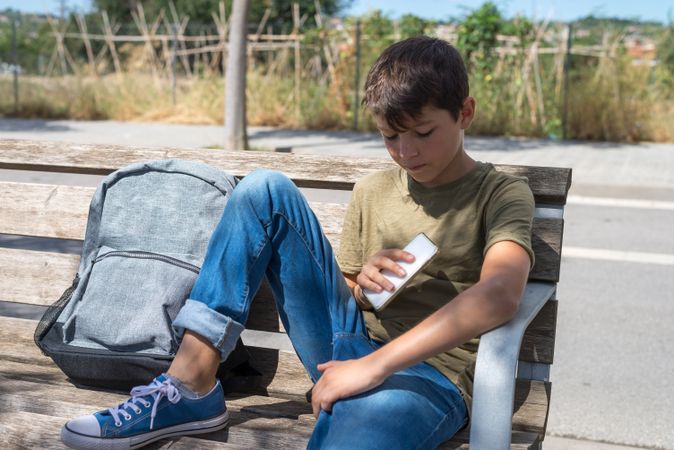Boy sitting on bench next to backpack holding phone