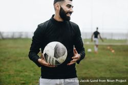 Smiling footballer standing on field holding a football during practice 0LKPRb