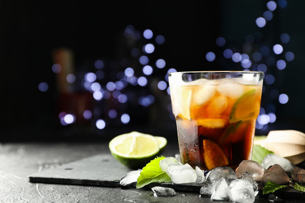 Glass with cola and lime slices, juicer, ice, mint against blurred lights background