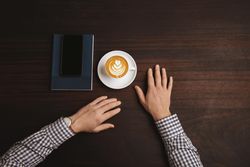 Top view of man in plaid with cappuccino, cell phone and notebook on wooden table