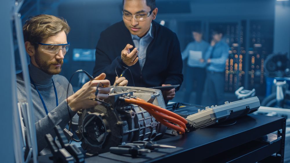 Two Professional Automotive Engineers with a Tablet Computer and Inspection Tools are Having a Conversation While Testing an Electric Engine in a High Tech Laboratory with a Concept Car Chassis.