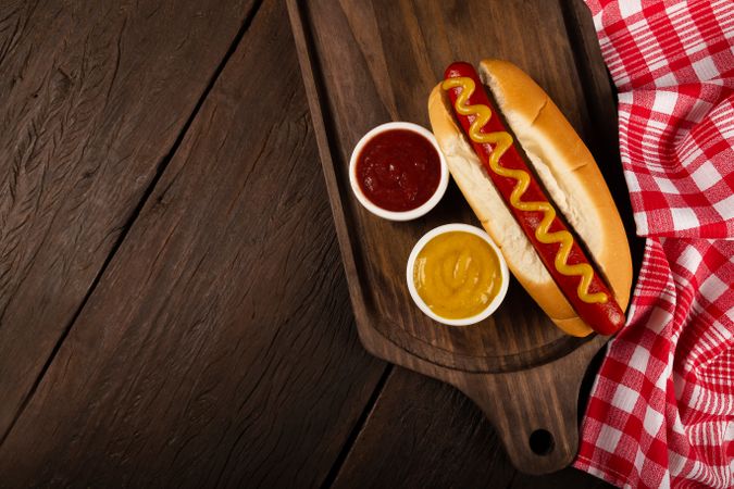 Top view of hot dog with mustard on wooden board with checkered dish towel and space for text