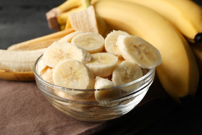 Bowl of banana slices, banana and towel on wooden background