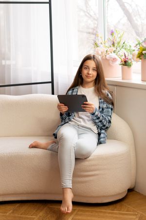 Teenage girl relaxing at home holding tablet near window.