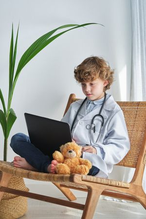 Boy doctor with toy bear and laptop on wicker chair
